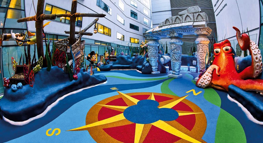 A colourful safe playground area in an enclosed outdoor space. The playground resembles an under the sea landscape with a sunken ship, a large octopus and a compass all made from Conica's wet pour